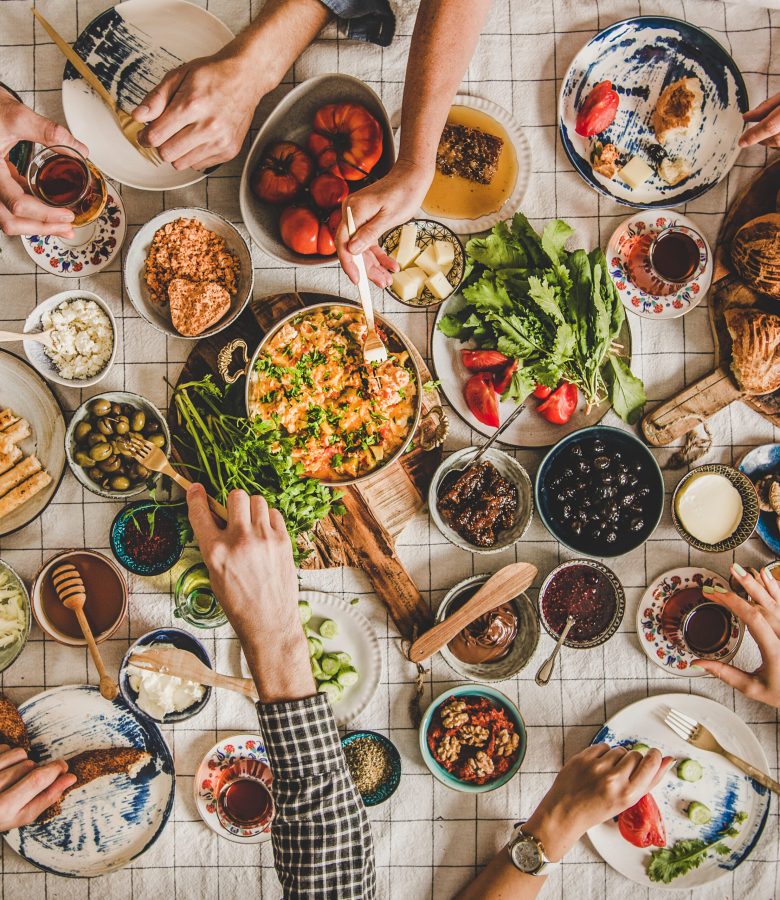 Flat-lay of peoples hands taking Turkish breakfast pastries, vegetables, greens, cheeses, fried eggs, jams, tea in tulip glasses over chekered linen tablecloth, top view, square crop