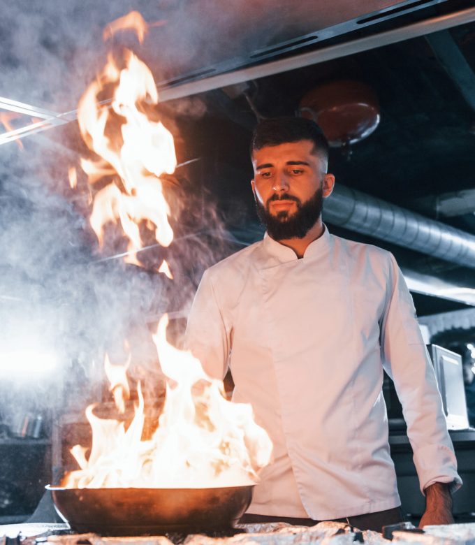 Uses frying pan. Chef in white uniform cooking food at kitchen. Busy day at work.